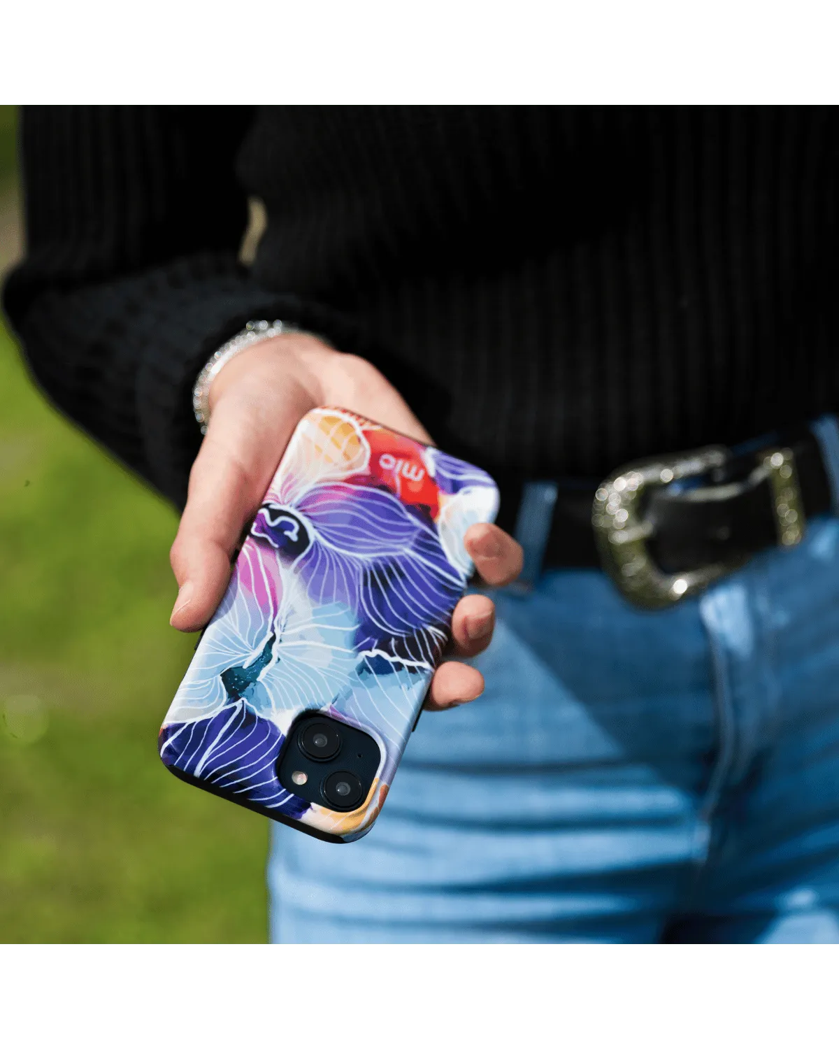 Hand houdt Samsung A55 5G met kleurrijk bloemenontwerphoesje van Mio vast in een buitenomgeving.